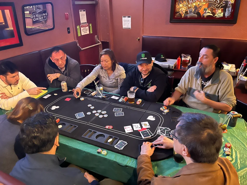 A group of men and women playing Texas Hold'em poker around a green felt table at a local pub social event.